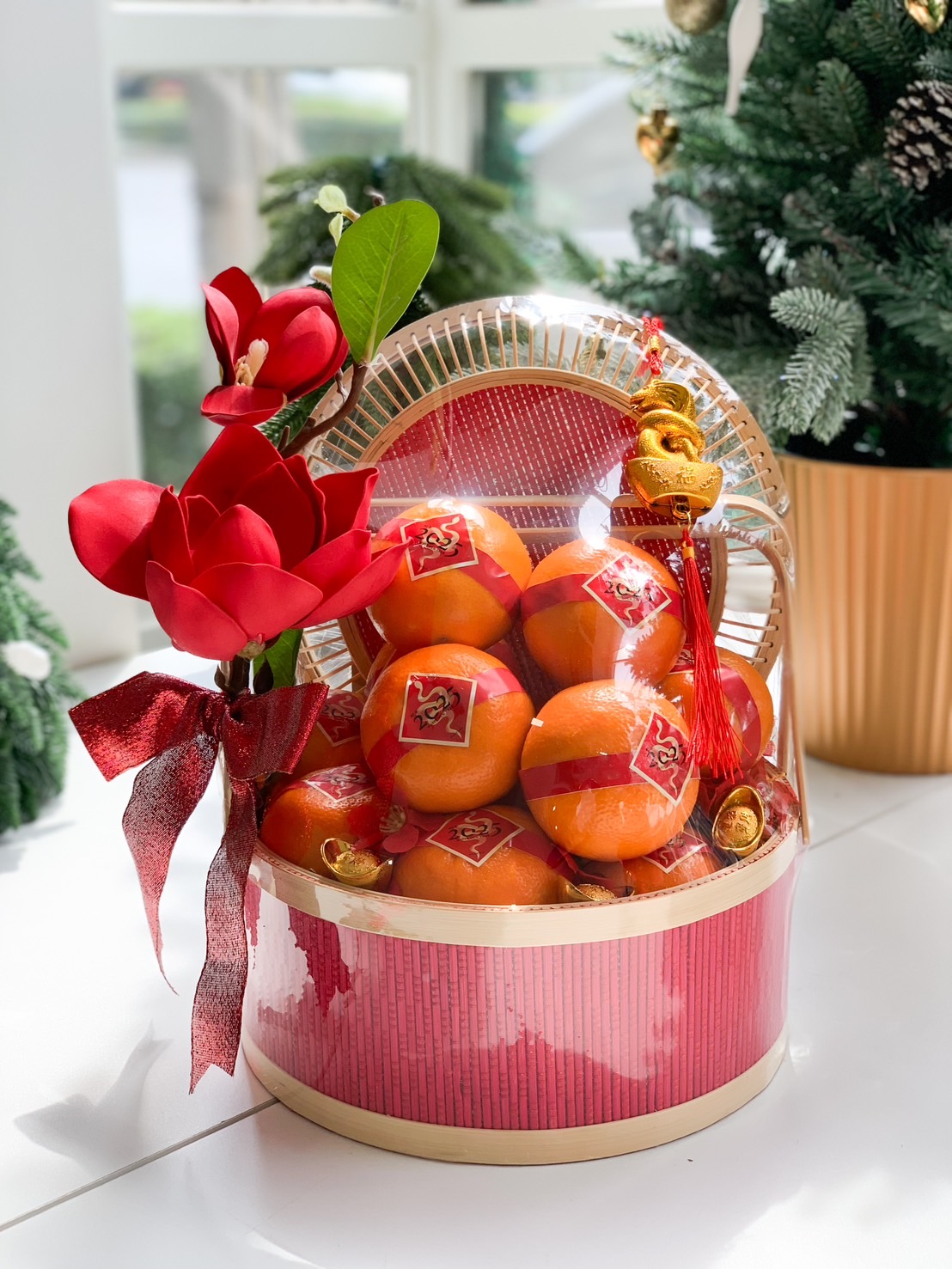 A festive Chinese New Year gift basket featuring fresh oranges wrapped with red ribbons, decorated with red flowers, gold ornaments, and a bamboo lid, presented in a vibrant red and gold design.