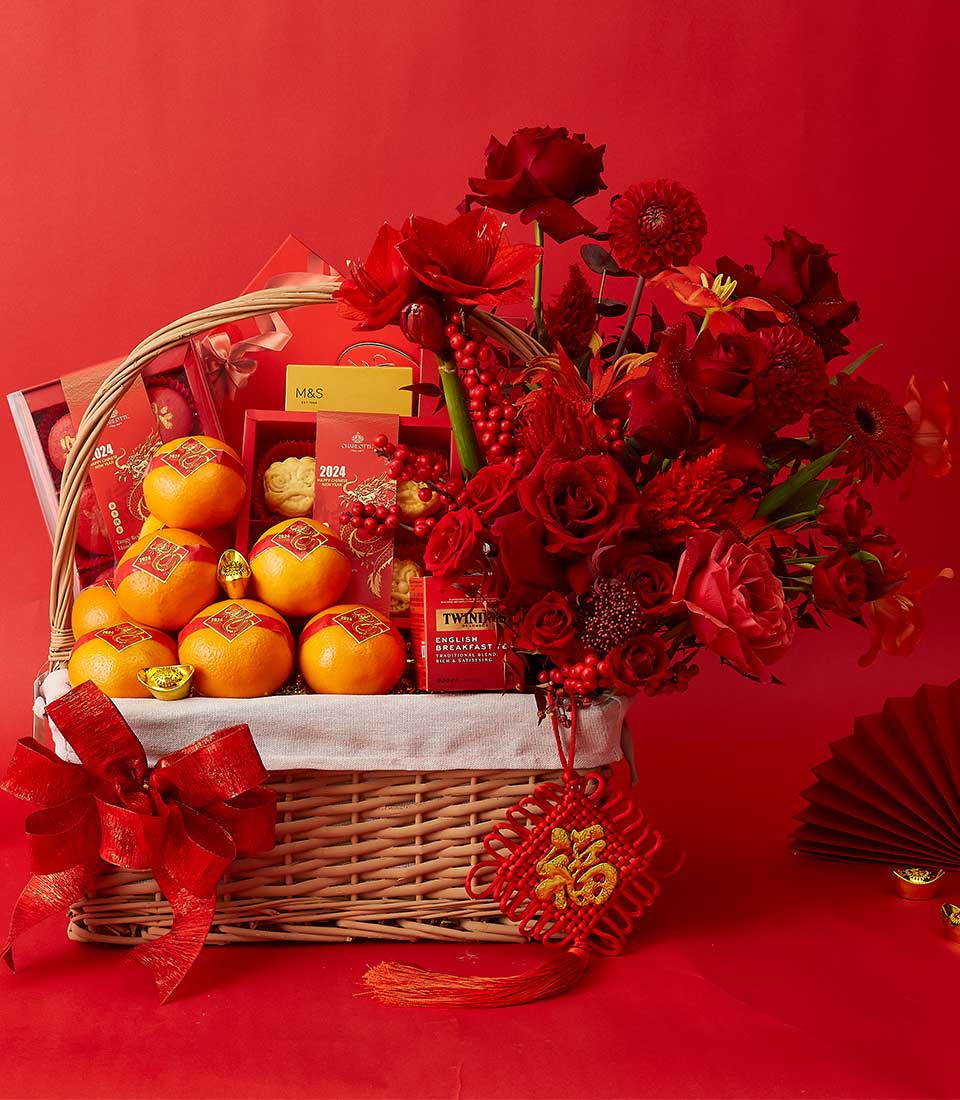 A festive gift basket sits against a red background. The basket contains oranges, gold-wrapped chocolates, and various red-packaged items. A lush arrangement of red flowers is attached to the basket. A red paper fan and Chinese ornament add to the festive decor.