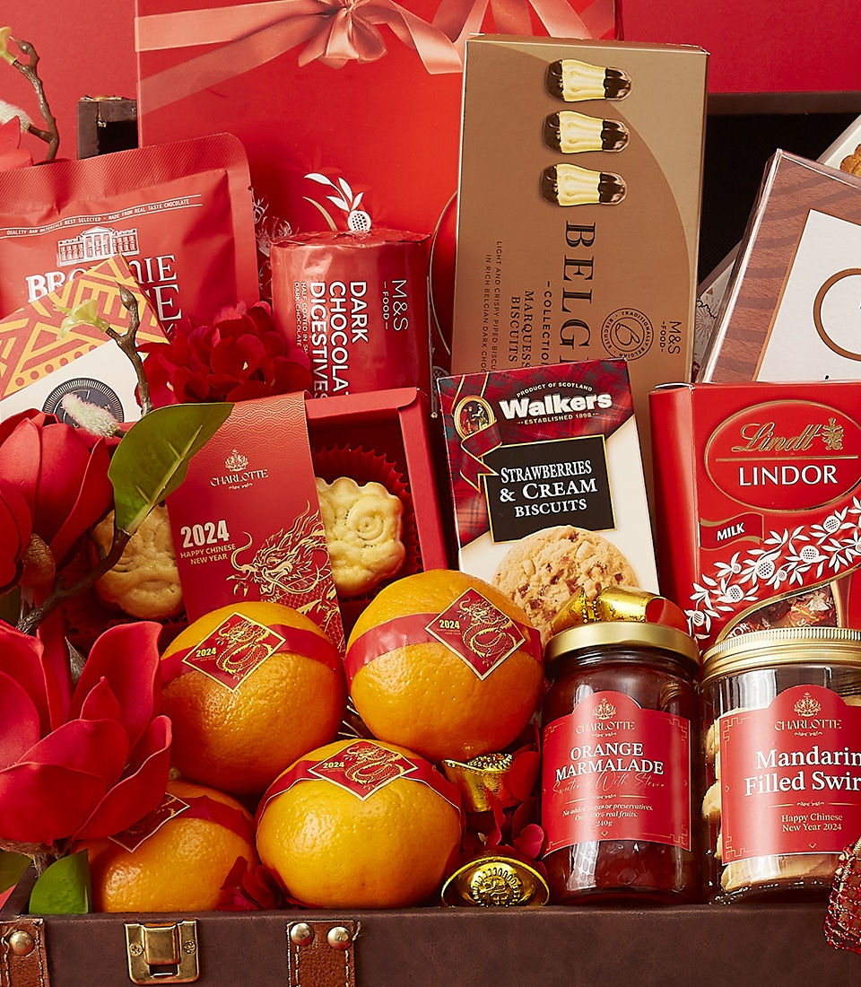 A festive gift basket filled with various items including oranges, strawberry and cream biscuits, orange marmalade, dark chocolate digestives, milk chocolate Lindor truffles, two Belgian chocolate bars, and a 2024 calendar. The items are arranged in a suitcase.