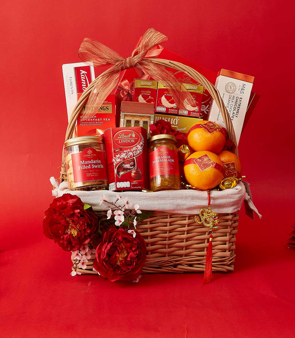 A wicker gift basket is filled with a variety of red-packaged goodies, including Lindt Lindor chocolates, Mandarin-filled Swiss chocolate, Ferrero Rocher, and more. The basket also contains fresh mandarins, red flowers, ribbons, and is set against a red background.