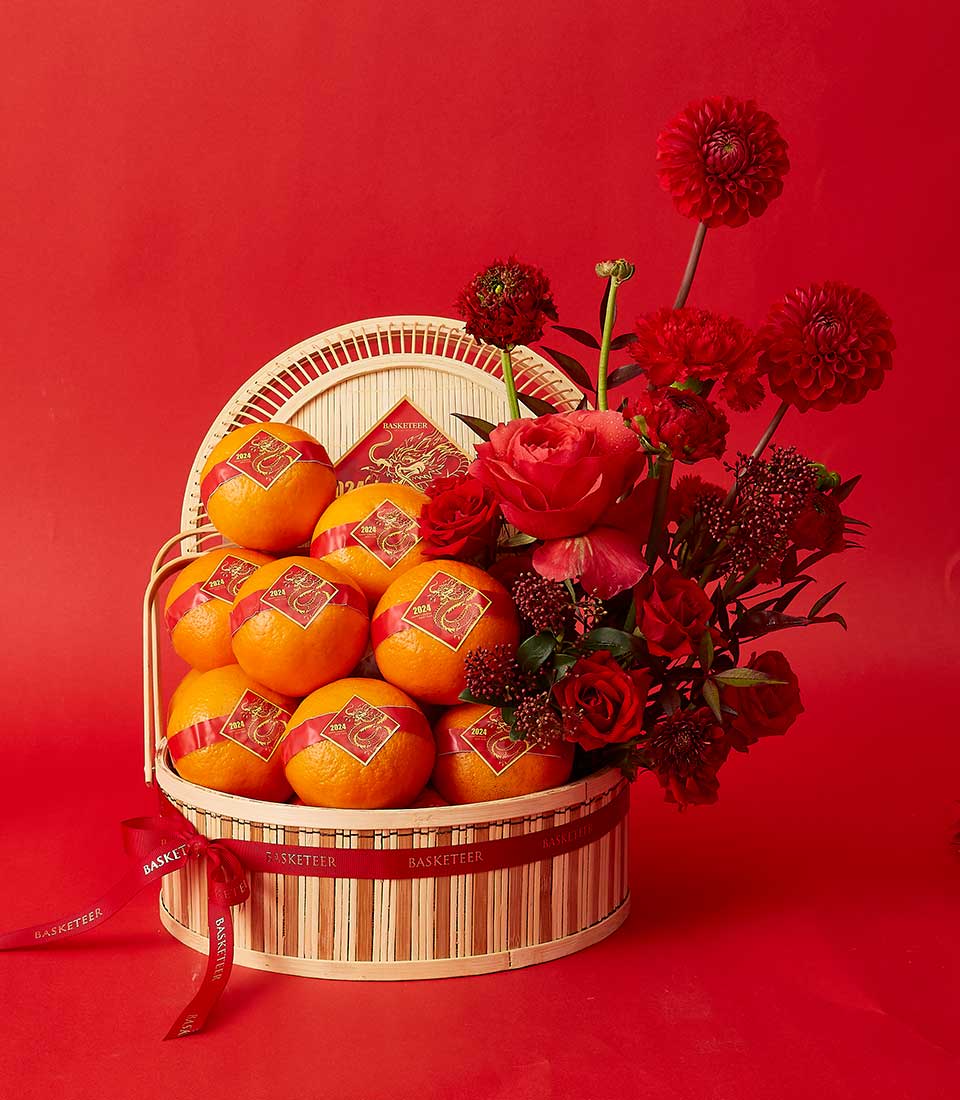 A bamboo basket is filled with several oranges adorned with red decorative stickers. The basket also contains a floral arrangement of vibrant red roses, dahlias, and other red flowers, all set against a solid red background.