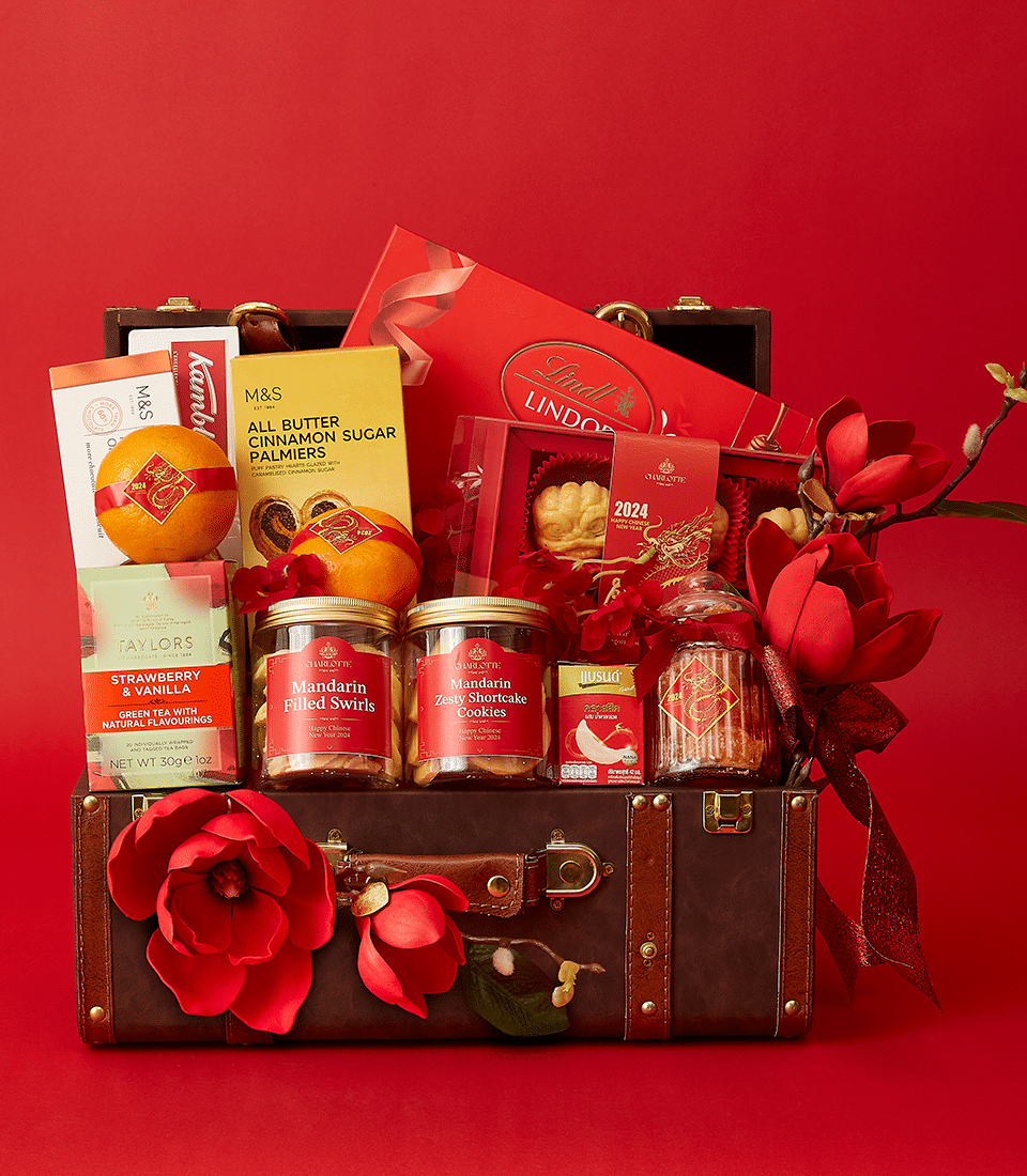 A festive gift hamper with a leather trunk filled with treats, including Lindt Lindor chocolates, M&S all-butter cinnamon sugar palmiers, Taylors Strawberry & Vanilla tea, Mandarin filled swirls, a pork pie, and a 2024 calendar, adorned with red flowers.