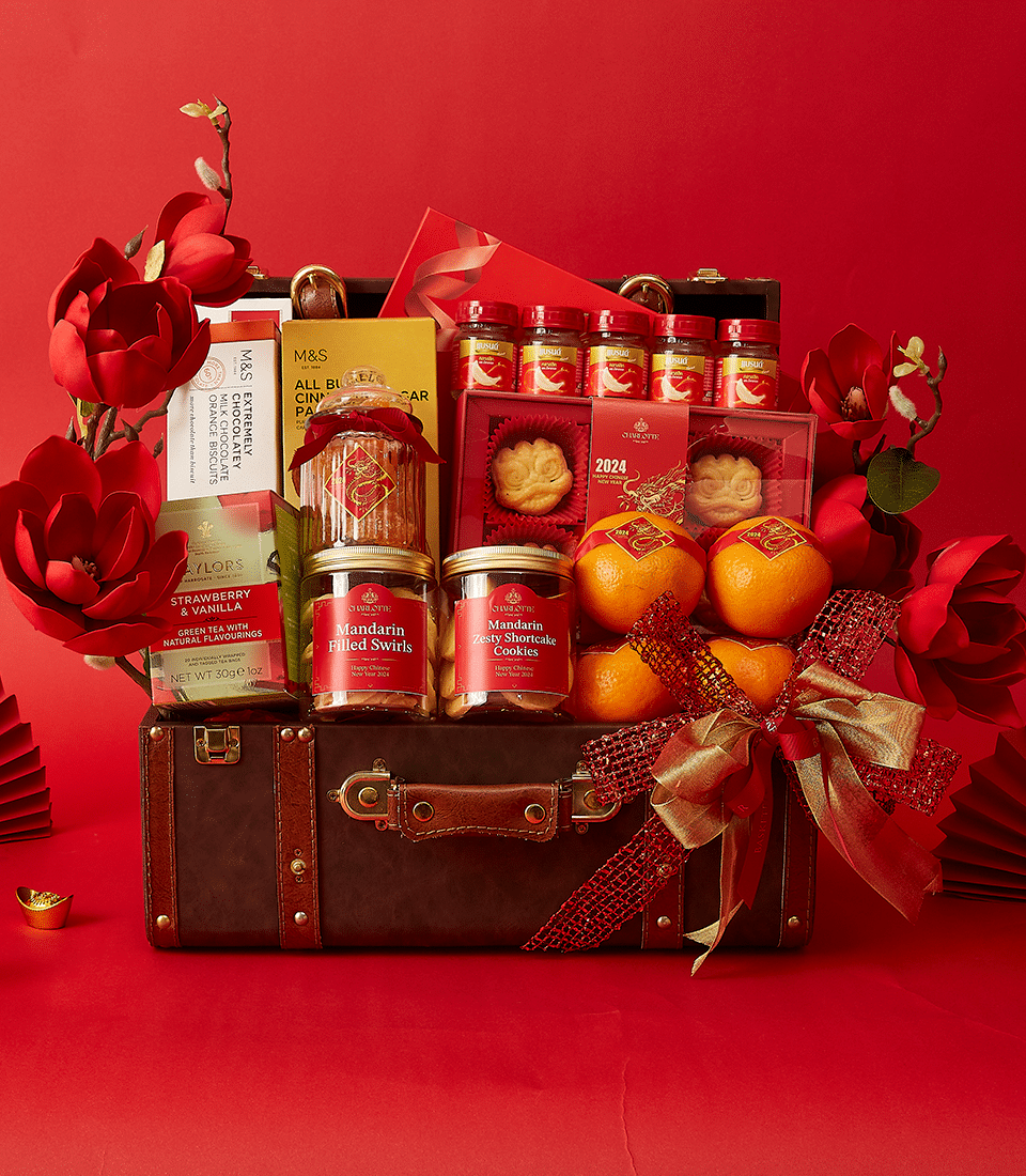 An ornate festive gift basket against a red background, featuring a brown suitcase adorned with a large red ribbon. The basket includes mandarin oranges, various sweets, cookies, teas, and decorative red and gold flowers, and gift containers.