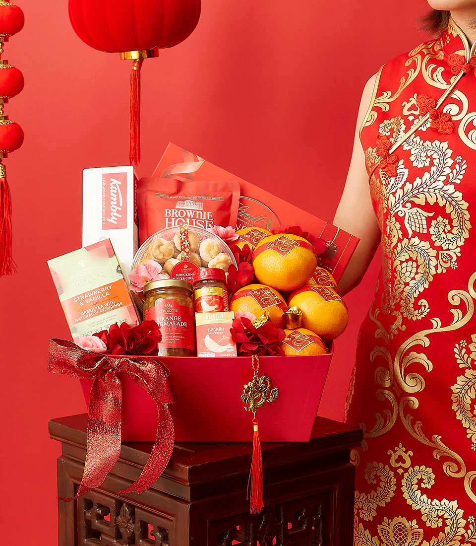 A woman in a red and gold traditional dress is standing beside a red gift basket containing oranges, snacks, tea, and other treats. Red lanterns hang in the background, highlighting the festive, celebratory atmosphere.