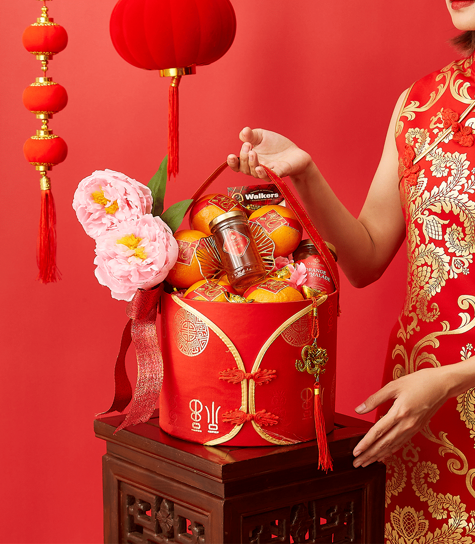 A person wearing a red and gold traditional outfit holds a festive red basket containing tangerines, a jar of Walker's candy, and adorned with pink flowers. The background features red lanterns and a decorative table, celebrating a festivity.