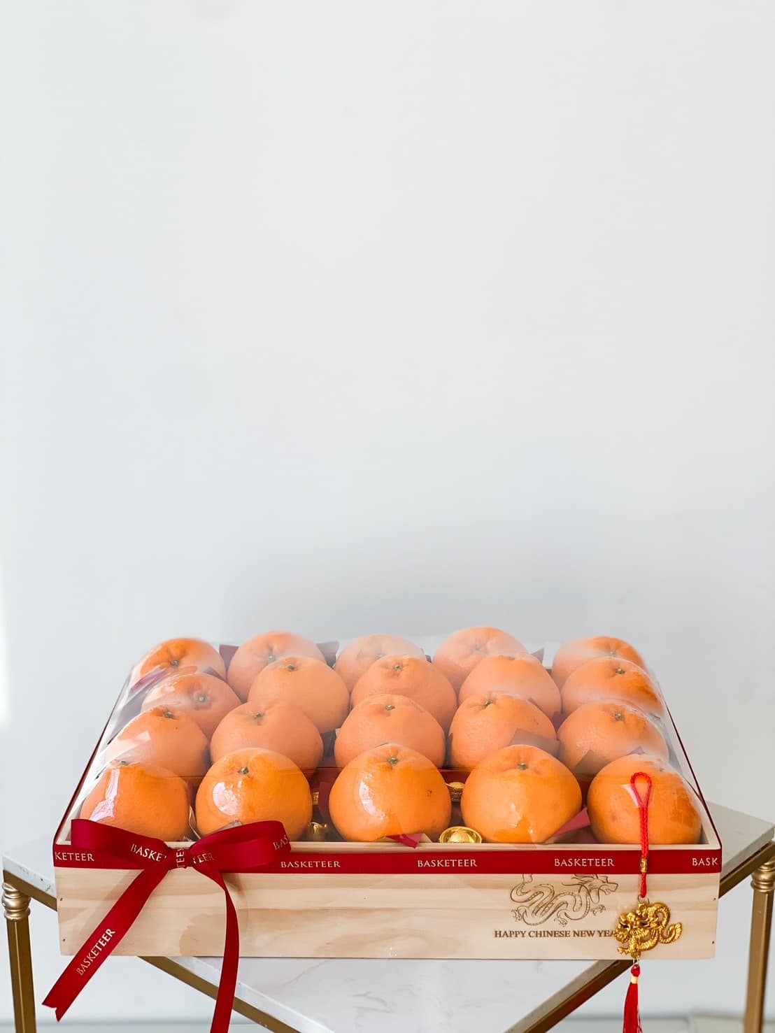 A wooden box holds neatly arranged persimmons wrapped in a transparent cover. The box is adorned with a red ribbon and a Chinese New Year decoration, featuring an intricate golden emblem. The background is plain white, making the vibrant colors stand out.