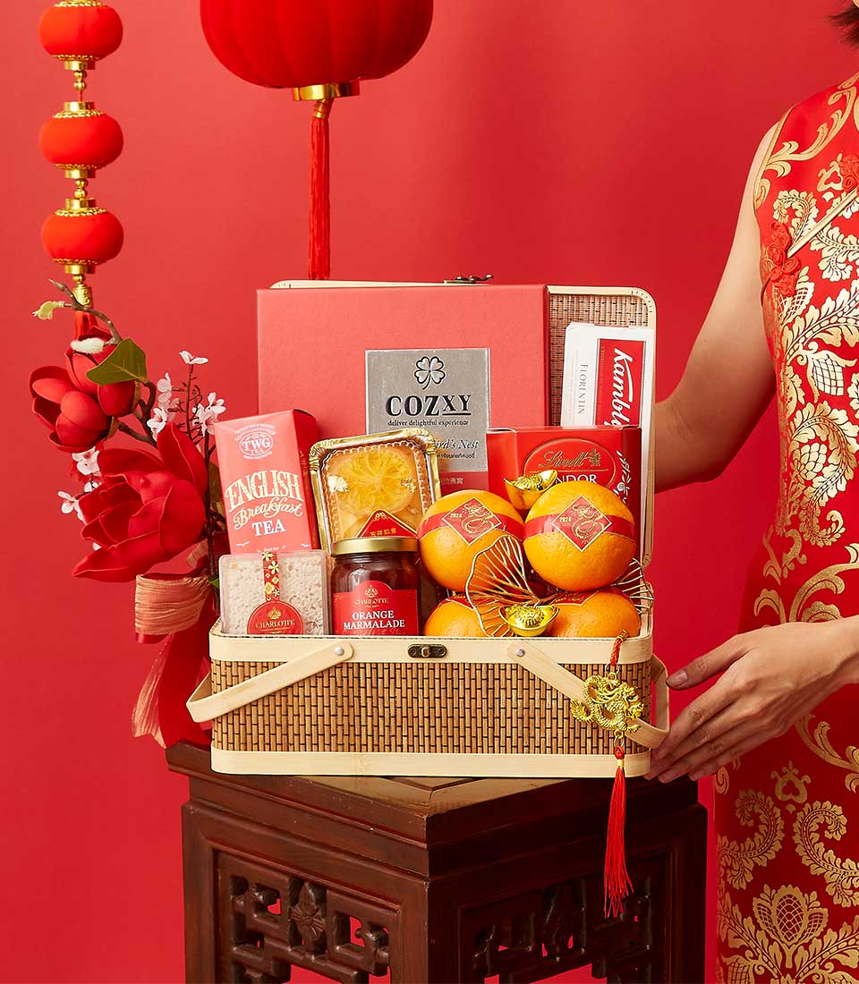 A festive gift basket on a wooden stand, filled with oranges, tea, cookies, and other treats. The basket also has decorative red envelopes and ornaments. It is set against a red background with a traditional lantern and a person in a patterned red dress holding it.