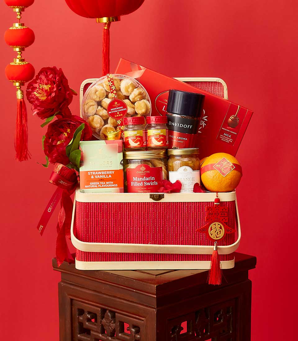 A festive red gift basket filled with assorted goodies, including shrimp rolls, mandarin-filled Swiss rolls, strawberry wafers, chocolates, and tea. The basket is adorned with red ribbons and placed on a wooden stand, with red lanterns hanging nearby.