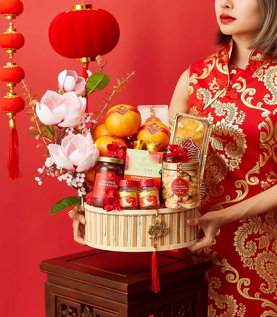 A person in a red and gold Chinese dress holds a festive basket containing flowers, oranges, crackers, tea, and jars of preserves. The background is red with hanging red lanterns, creating a celebratory atmosphere.