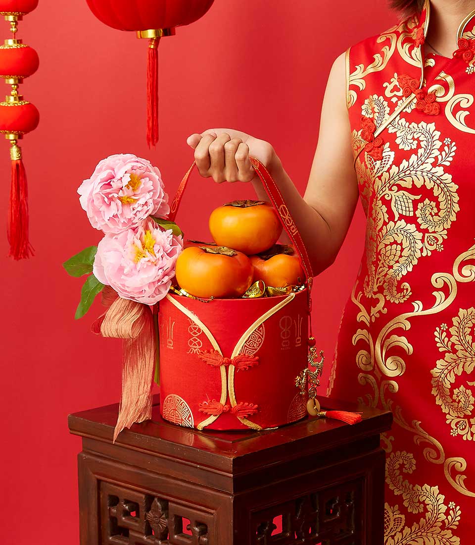 A person in a red and gold traditional Chinese outfit is holding a red basket adorned with gold designs, containing persimmons and pink flowers. The setting includes red lanterns and a red background, evoking a festive atmosphere.
