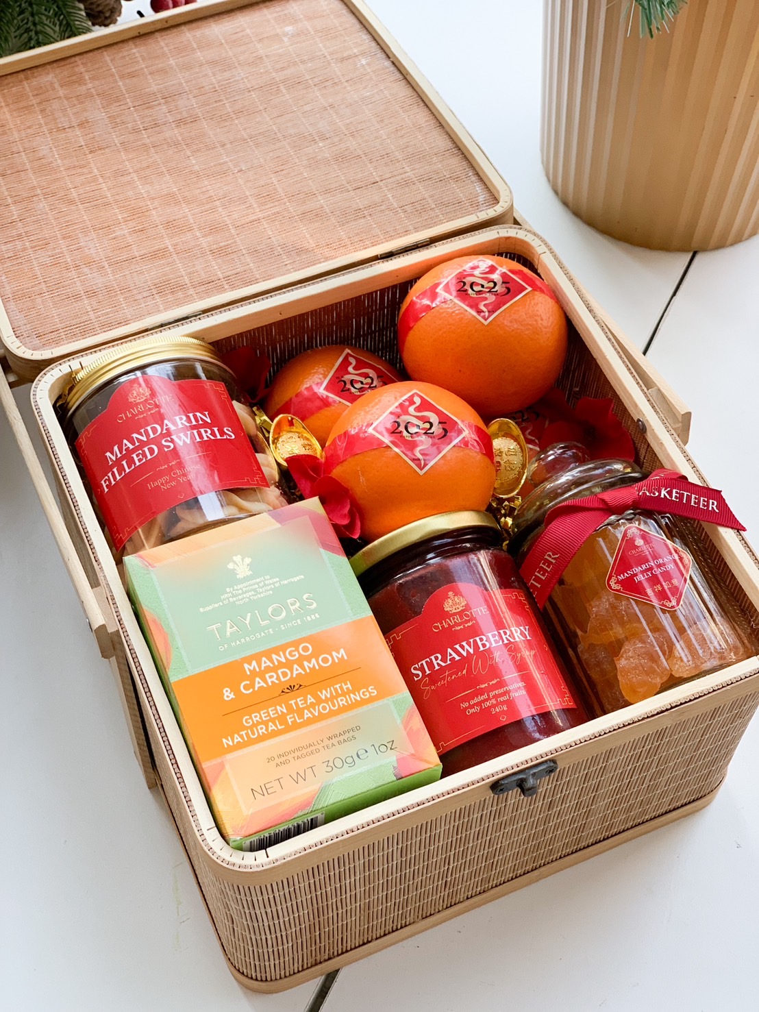 Elegant Chinese New Year gift basket featuring fresh oranges, mango cardamom tea, strawberry jam, mandarin candies, and festive decorations in a bamboo-style box.