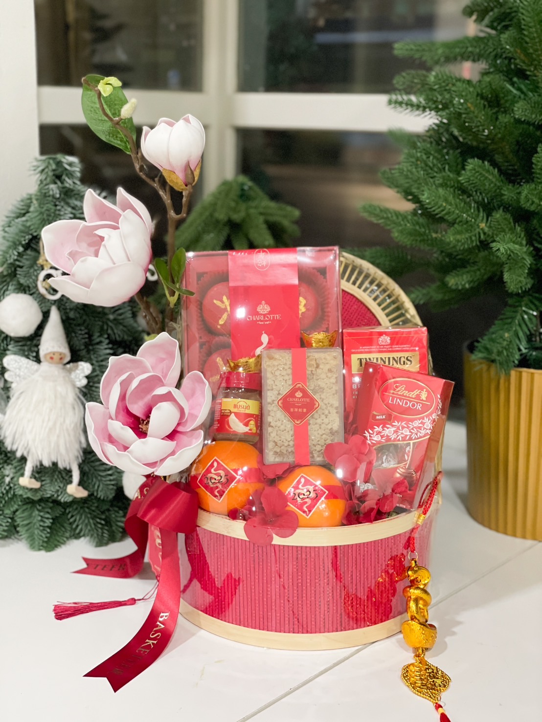 A stunning gift basket containing mandarin oranges, premium chocolates, Twinings tea, and festive snacks, elegantly adorned with artificial magnolias and vibrant red and gold decorations.