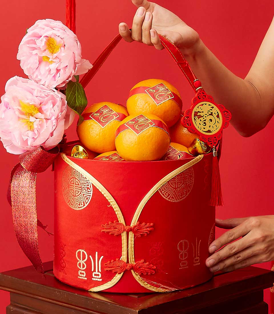 A decorative red basket filled with oranges decorated with Chinese New Year symbols and wrapped in red and gold paper. The basket is adorned with artificial pink flowers, a red ribbon, and a hanging gold and red ornament. A person's hand is touching the basket.