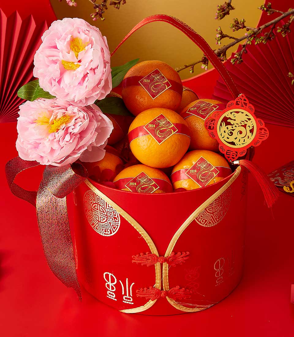 A red decorative bucket filled with oranges, each adorned with a square red and gold design, symbolizing prosperity. Two pink peonies and a red ribbon with a round ornament enhance the festive presentation. Red fans and branches with small blossoms are in the background.