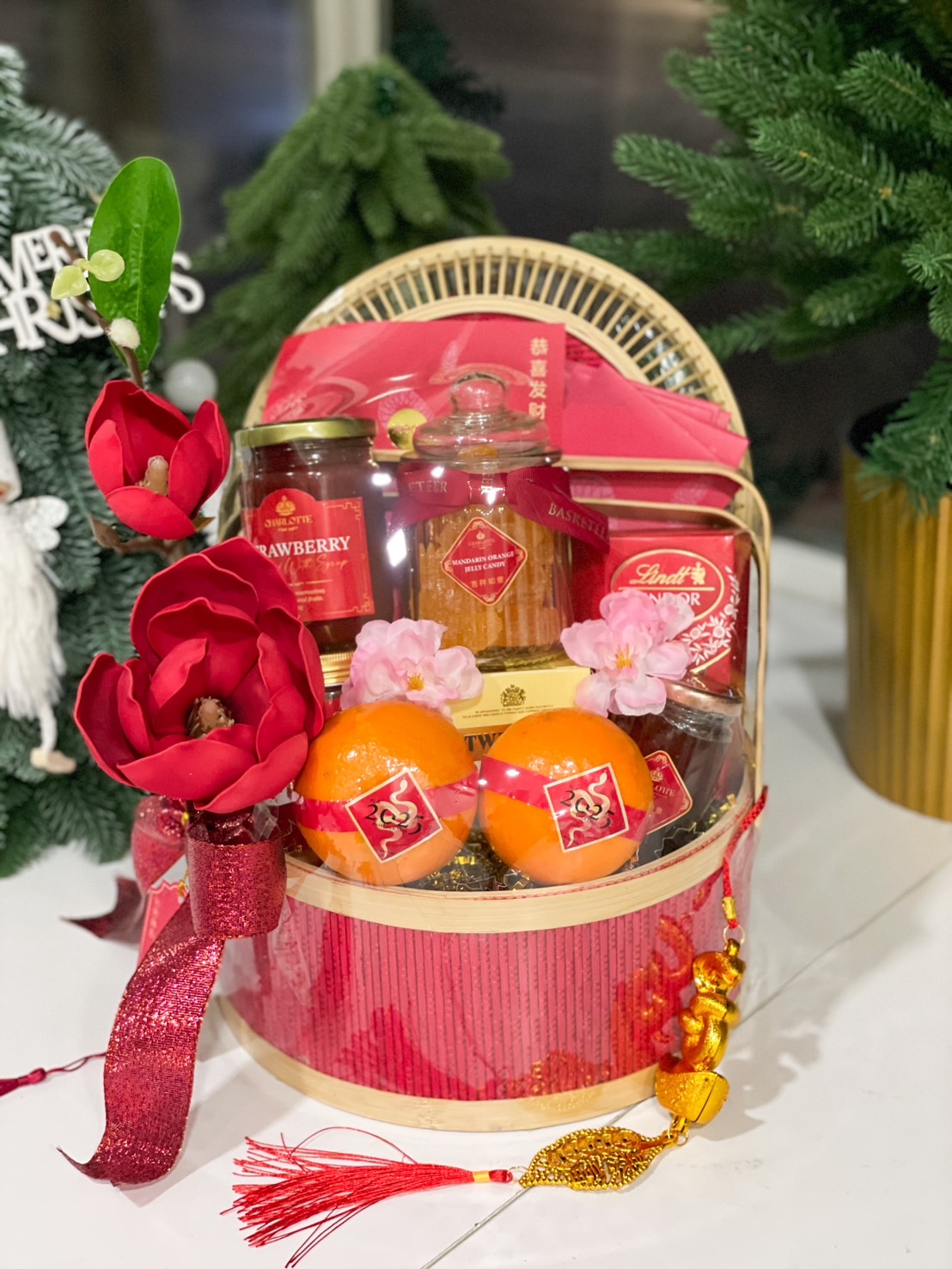 A festive gift basket containing various items like Lindor chocolates, jars, oranges, and a decorative glass jar. The basket has a natural woven texture, and a red magnolia flower arrangement adorns the left side, creating a vibrant and elegant display.
