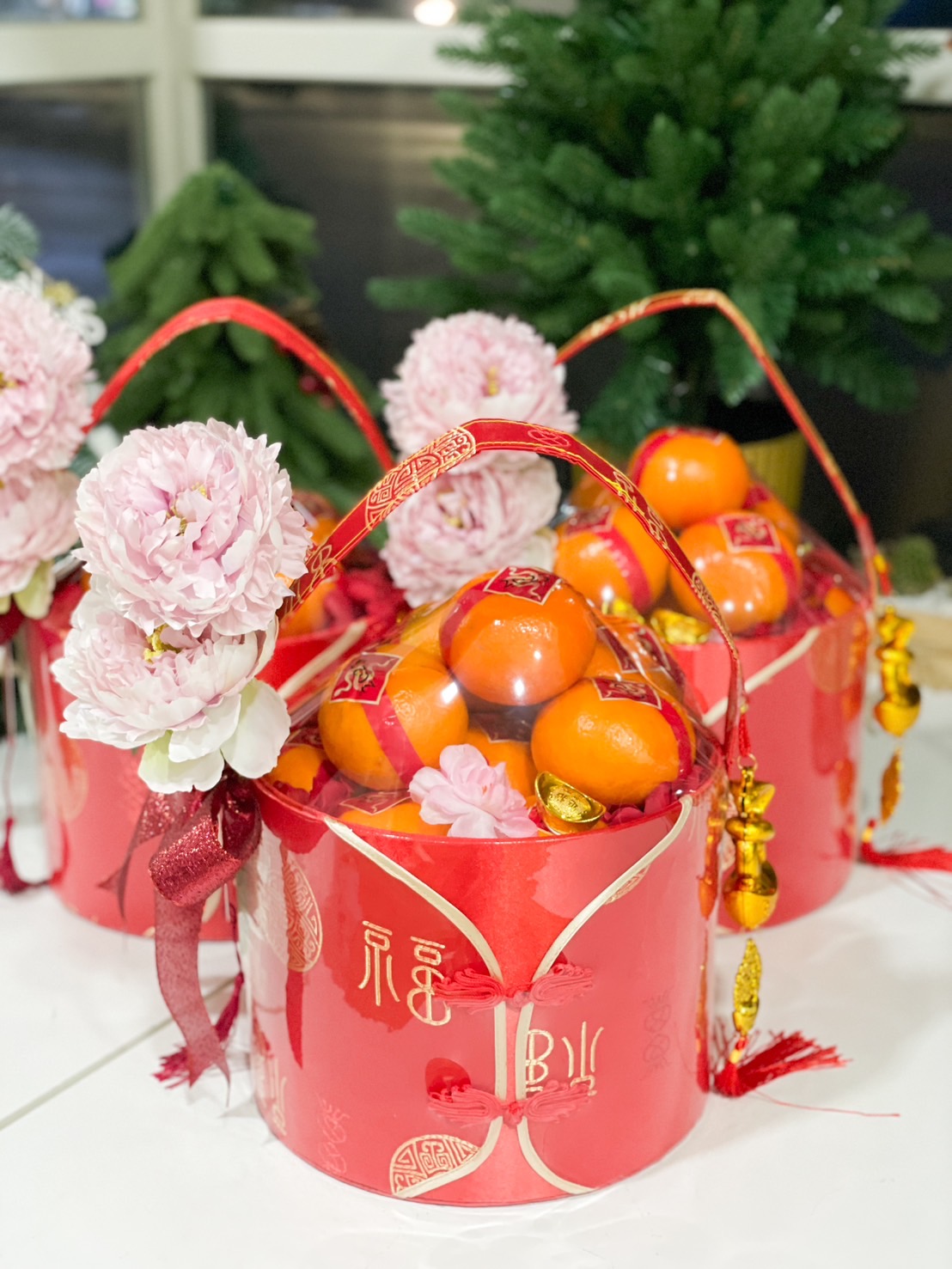 Gorgeous Chinese New Year hampers featuring pink peonies, fresh oranges, and vibrant red decorative buckets adorned with golden motifs, perfect for festive gifting.