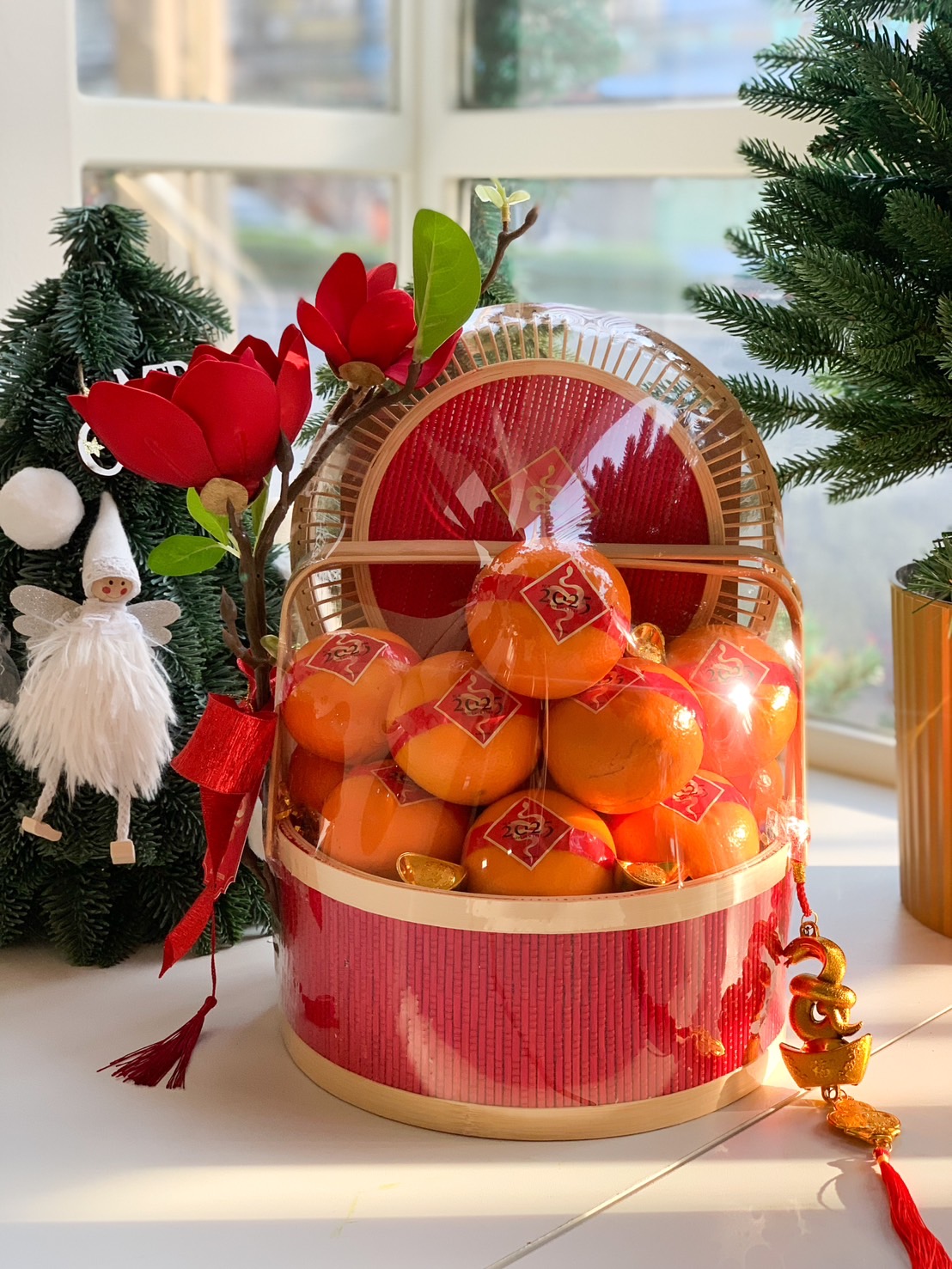 A festive Chinese New Year gift basket featuring fresh oranges wrapped with red ribbons, decorated with red flowers, gold ornaments, and a bamboo lid, presented in a vibrant red and gold design.