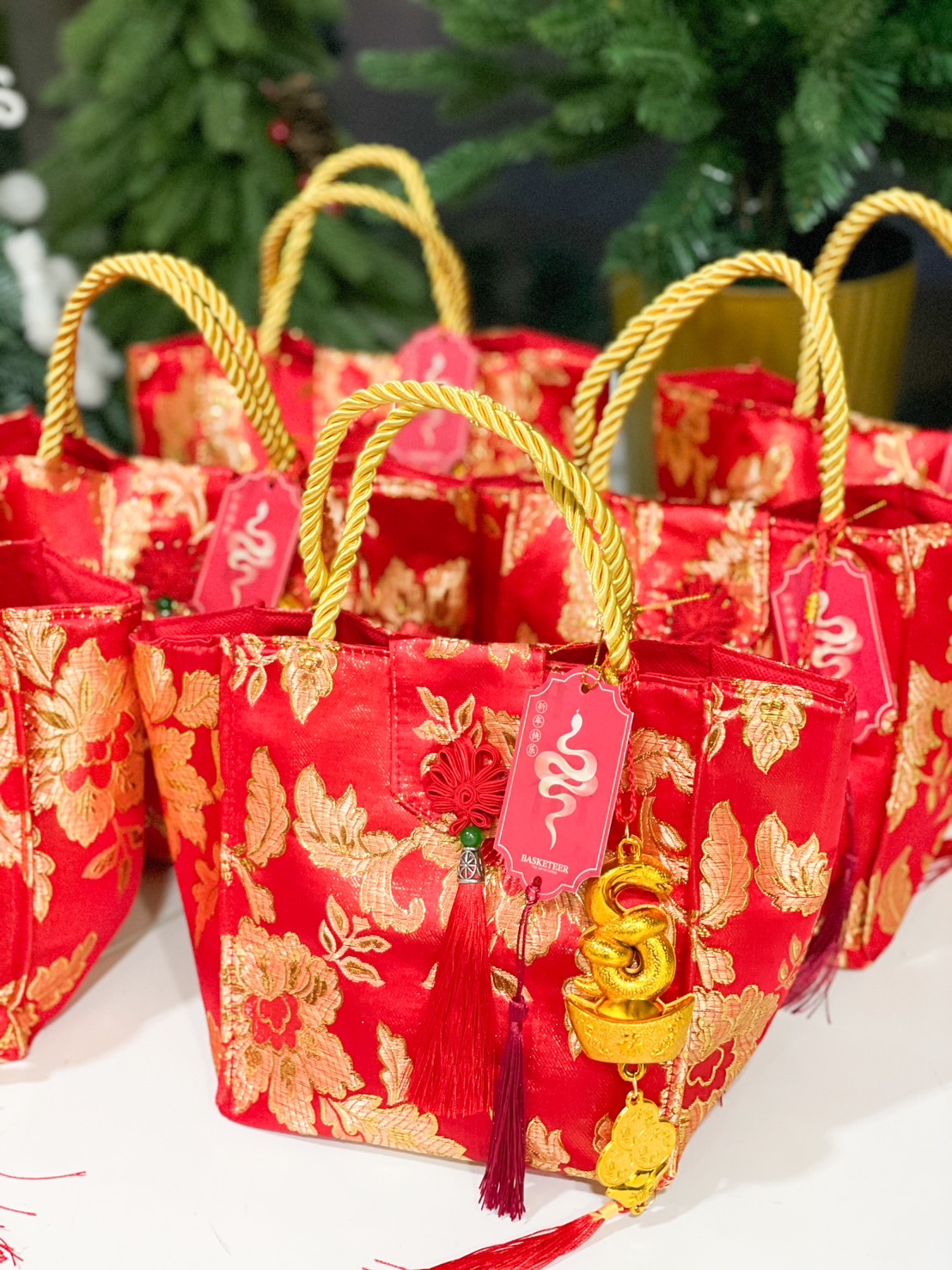 Close-up of a Chinese New Year festive tote bags in red and gold floral brocade, accented with gold rope handles, red tassels, and golden snake charms.
