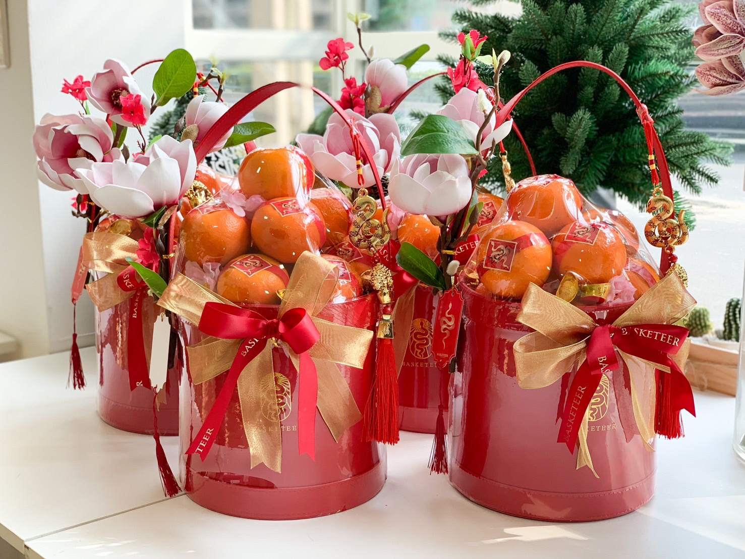 Red Chinese New Year gift basket with mandarin oranges, gold charms, and decorative blossoms, tied with festive ribbons.