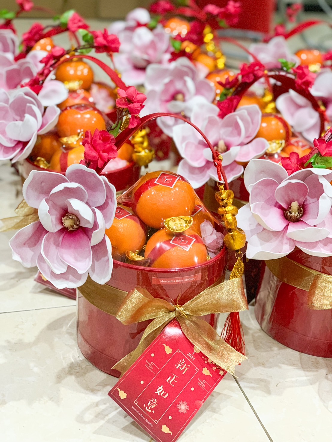 Red premium Chinese New Year gift basket featuring fresh oranges, pink decorative flowers, a golden bow, and festive gold accents, perfect for celebrations.