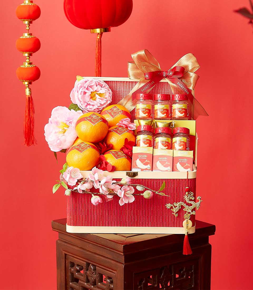 A festive red gift box adorned with pink and white flowers, containing oranges, bottled drinks, and small packets. The box is topped with a gold bow and placed on a decorative wooden stand, accompanied by hanging red lanterns in the background.