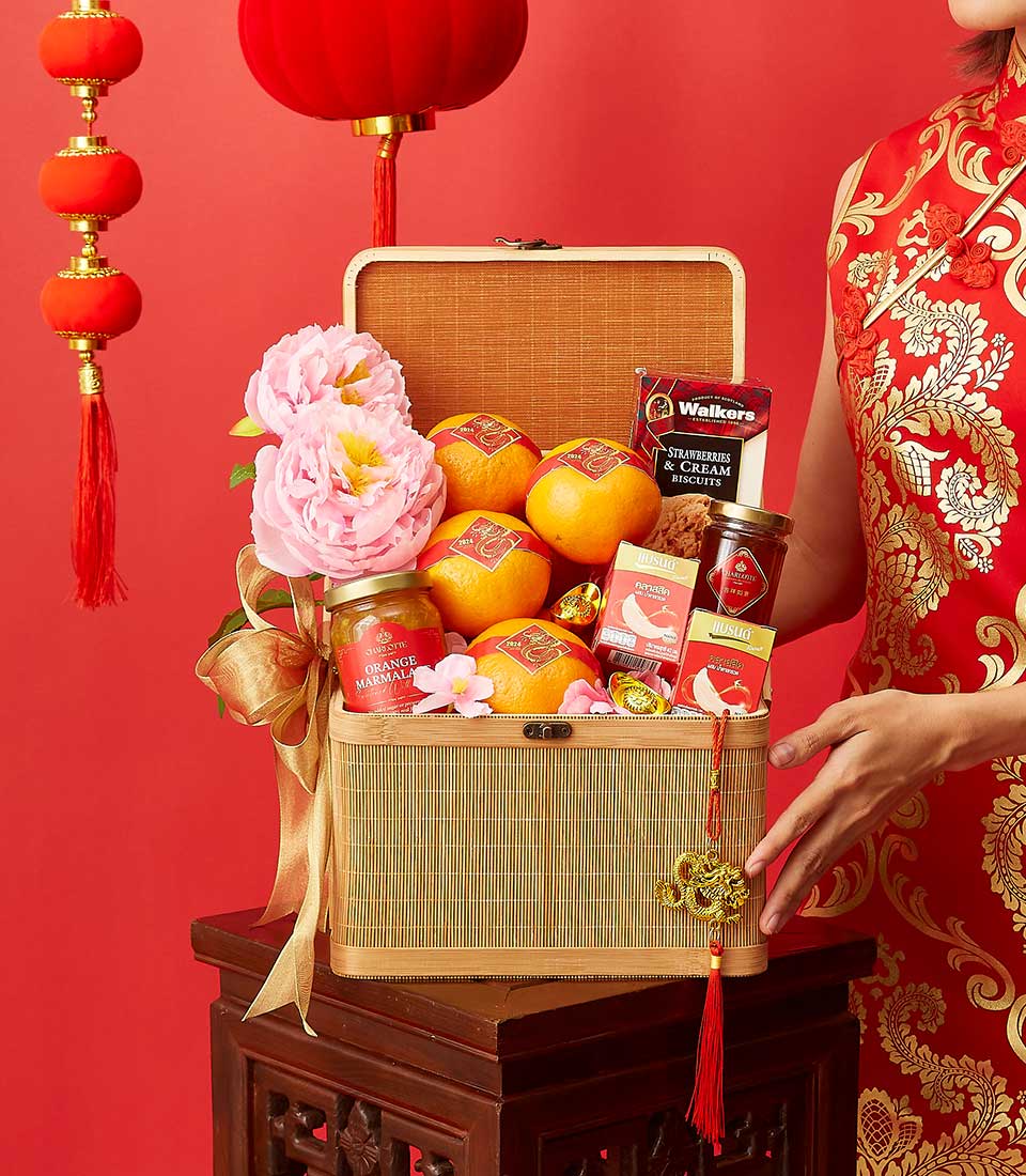 A person in a red floral dress presents an open woven basket filled with oranges, decorative flowers, honey, tea, and cookies. The background is red, adorned with hanging red lanterns and tassels, creating a festive atmosphere.