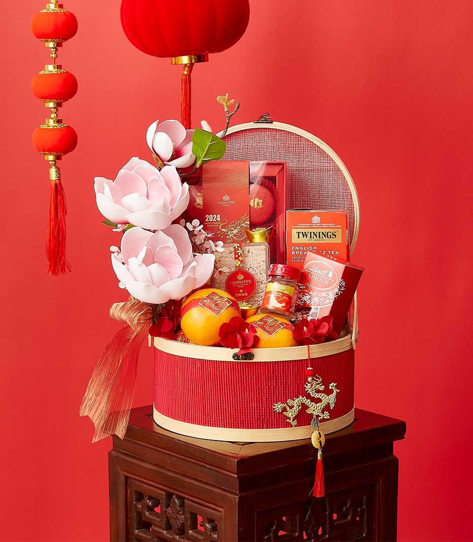 A festive red and gold basket sits on a wooden stand, filled with flowers, oranges, tea, and various gift items. It is decorated with a ribbon, surrounded by hanging red lanterns, all set against a vibrant red background to celebrate a traditional festival.