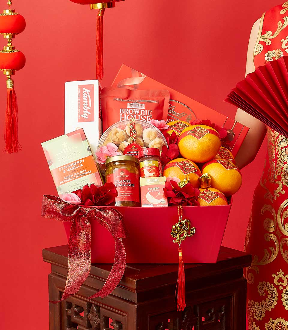A red gift basket features a variety of items including oranges, a candle, a fan, cookies, tea, and sweets. The basket is decorated with red and gold ribbons and Chinese charms. Two red lanterns hang in the background, and a person in red traditional attire stands beside it.