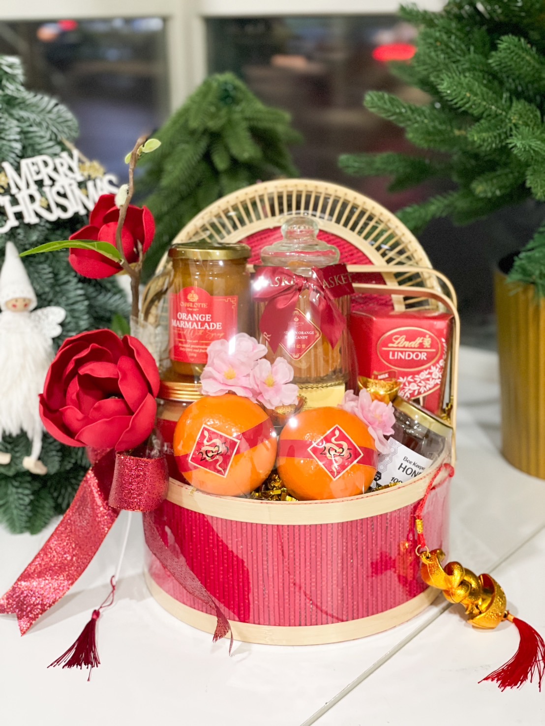 A festive gift basket containing various items like Lindor chocolates, jars, oranges, and a decorative glass jar. The basket has a natural woven texture, and a red magnolia flower arrangement adorns the left side, creating a vibrant and elegant display.