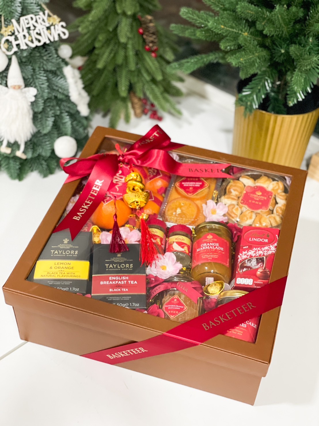 A festive gift box filled with assorted fruits, chocolates, cookies, and preserves. The box includes oranges, a fruitcake, Lindt Lindor chocolates, jars of mango, mandarin, guava preserves, strawberry and passionfruit preserves, and crunchy cookies. The items are decorated with red packaging and pink flowers.