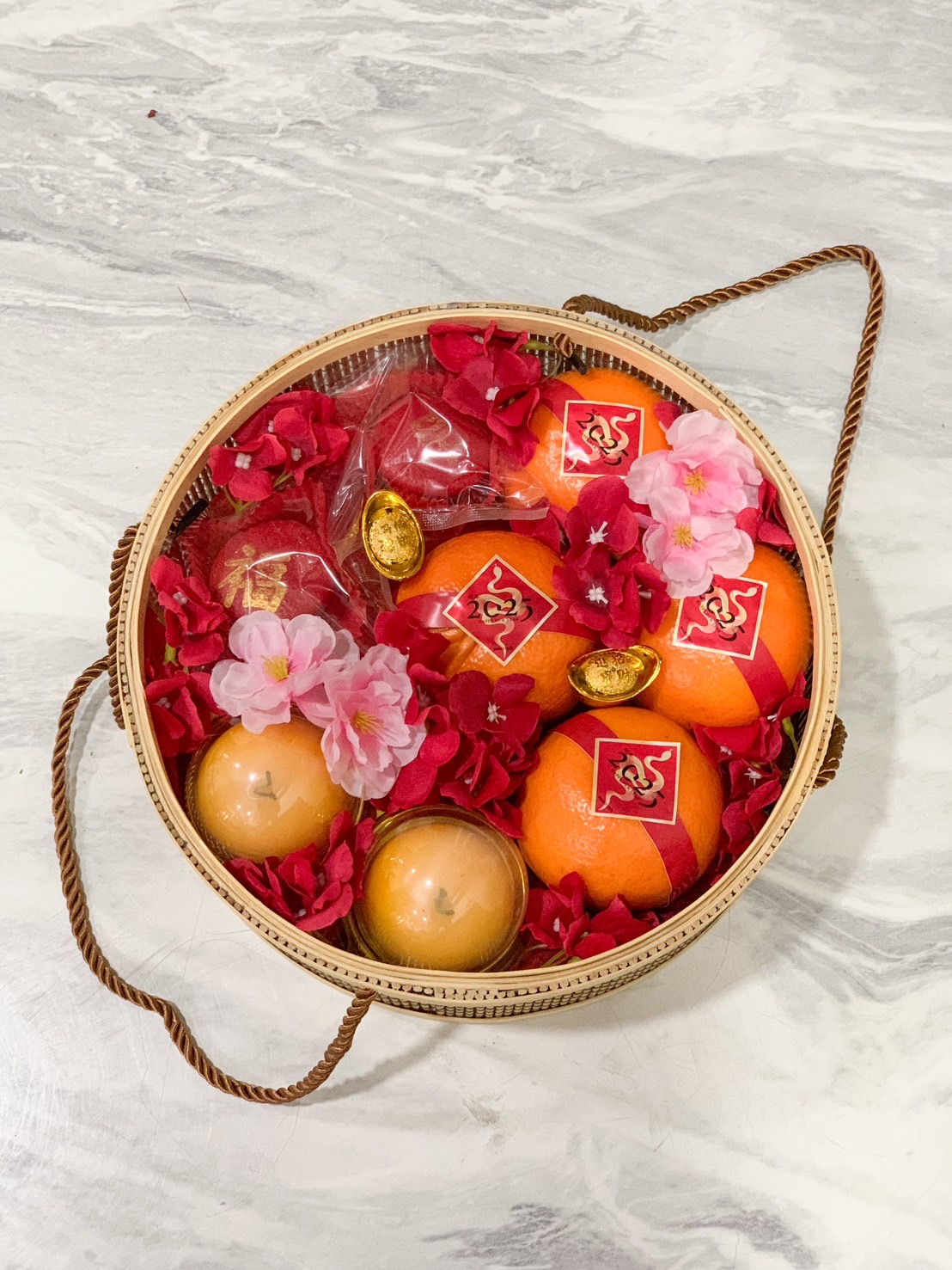 Bamboo round gift basket with Chinese New Year oranges, red flowers, candles, and gold charms, celebrating the Year of the Snake.