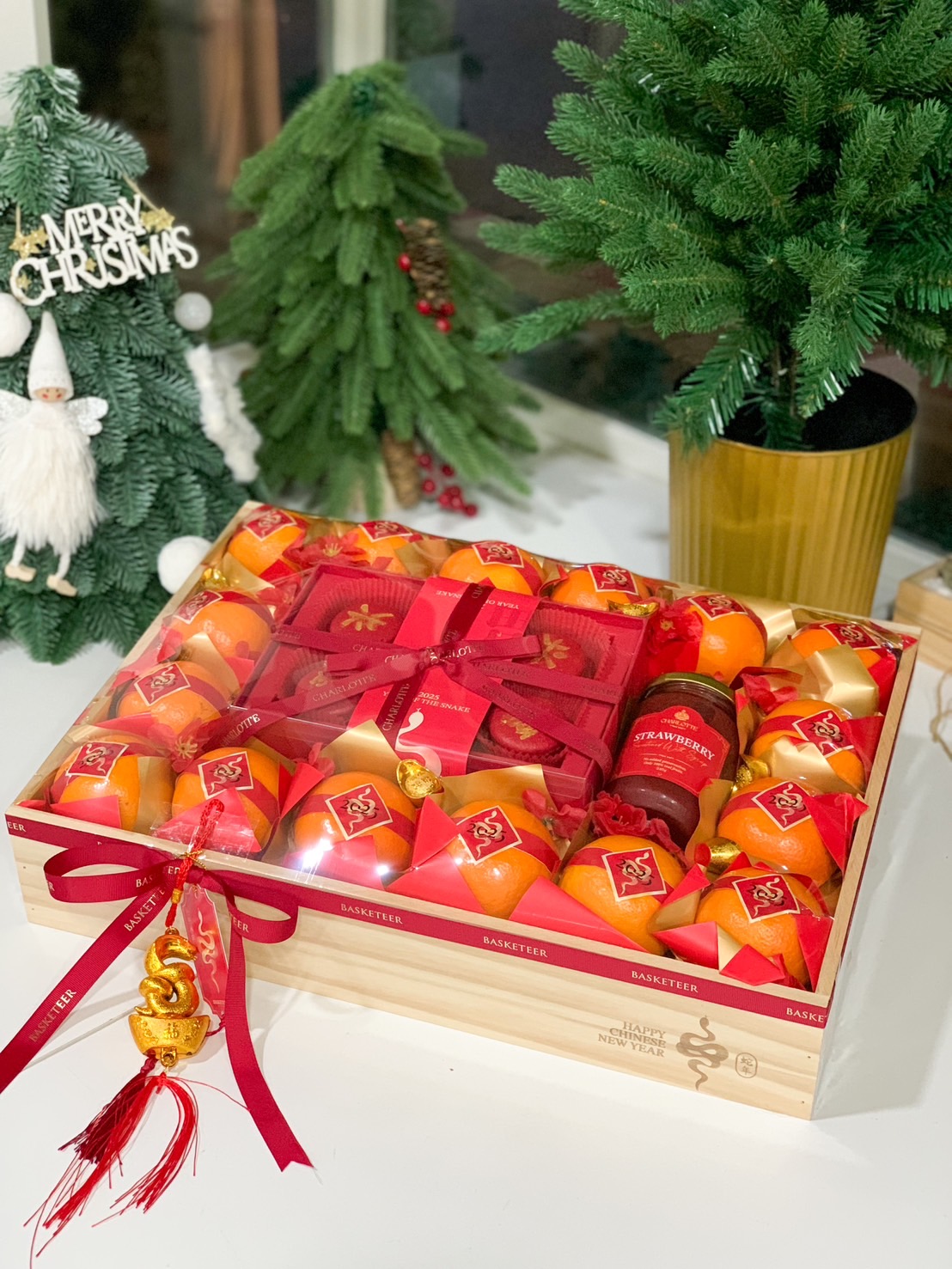 A festively decorated wooden box filled with vibrant oranges wrapped in red paper, several red envelopes, a jar of preserved fruits, and a decorative tassel, all arranged against a red and gold background, celebrating the Chinese New Year.