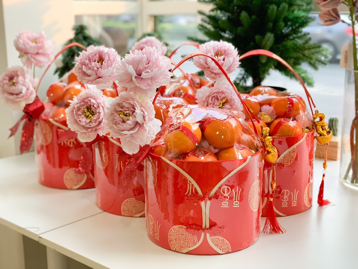 Gorgeous Chinese New Year hampers featuring pink peonies, fresh oranges, and vibrant red decorative buckets adorned with golden motifs, perfect for festive gifting.
