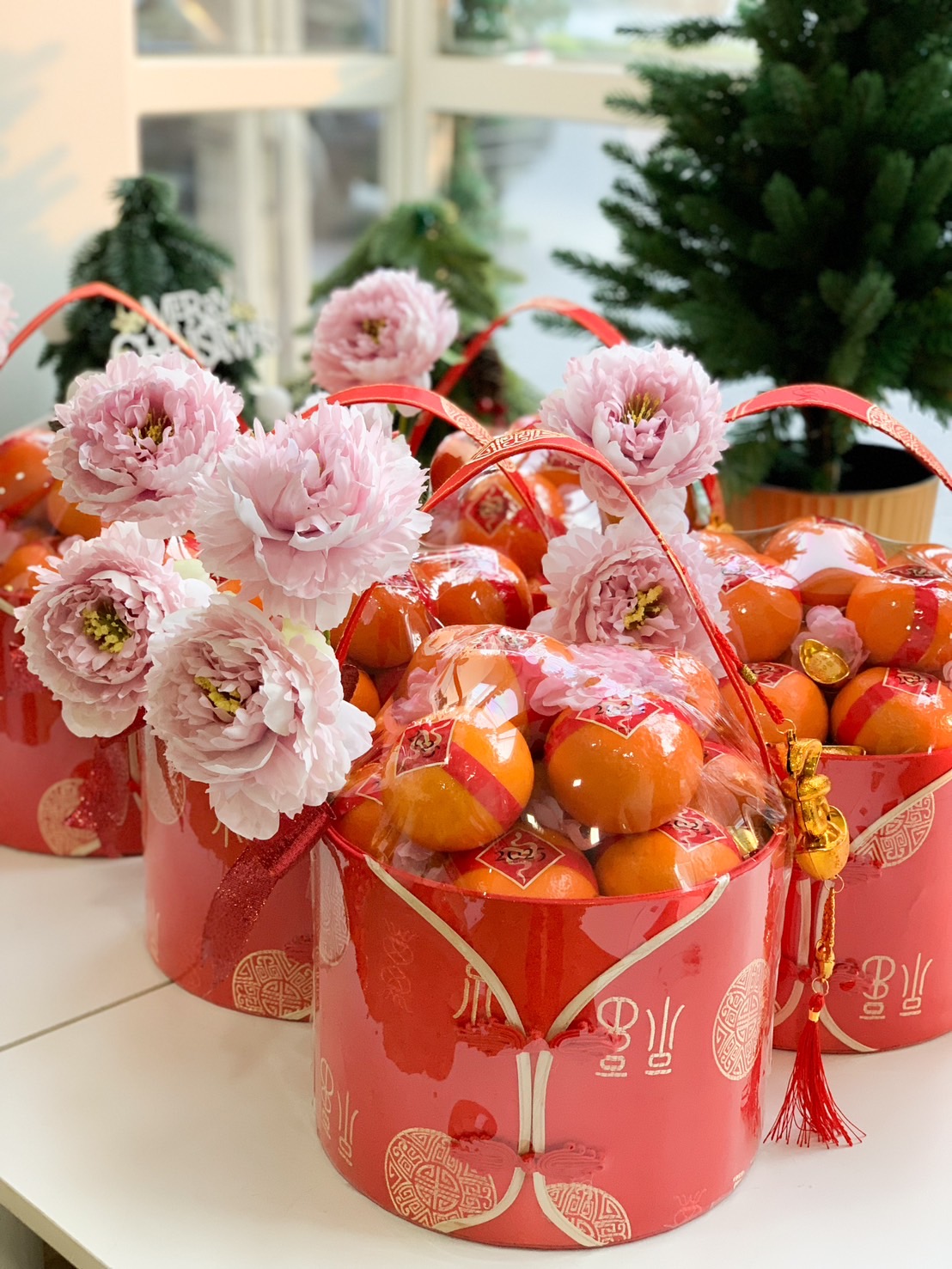 Close-up of a Gorgeous Chinese New Year hampers featuring pink peonies, fresh oranges, and vibrant red decorative buckets adorned with golden motifs, perfect for festive gifting.