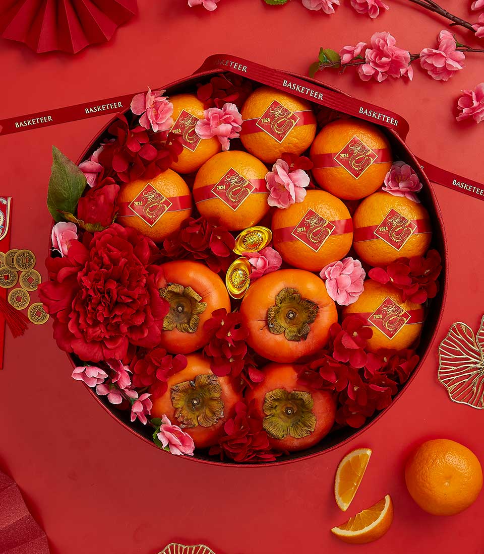 A festive arrangement featuring oranges adorned with red and gold stickers and five persimmons, surrounded by vibrant red flowers and pink blossoms. The setup is on a red background with scattered fan-shaped paper decorations and a red ribbon labeled 