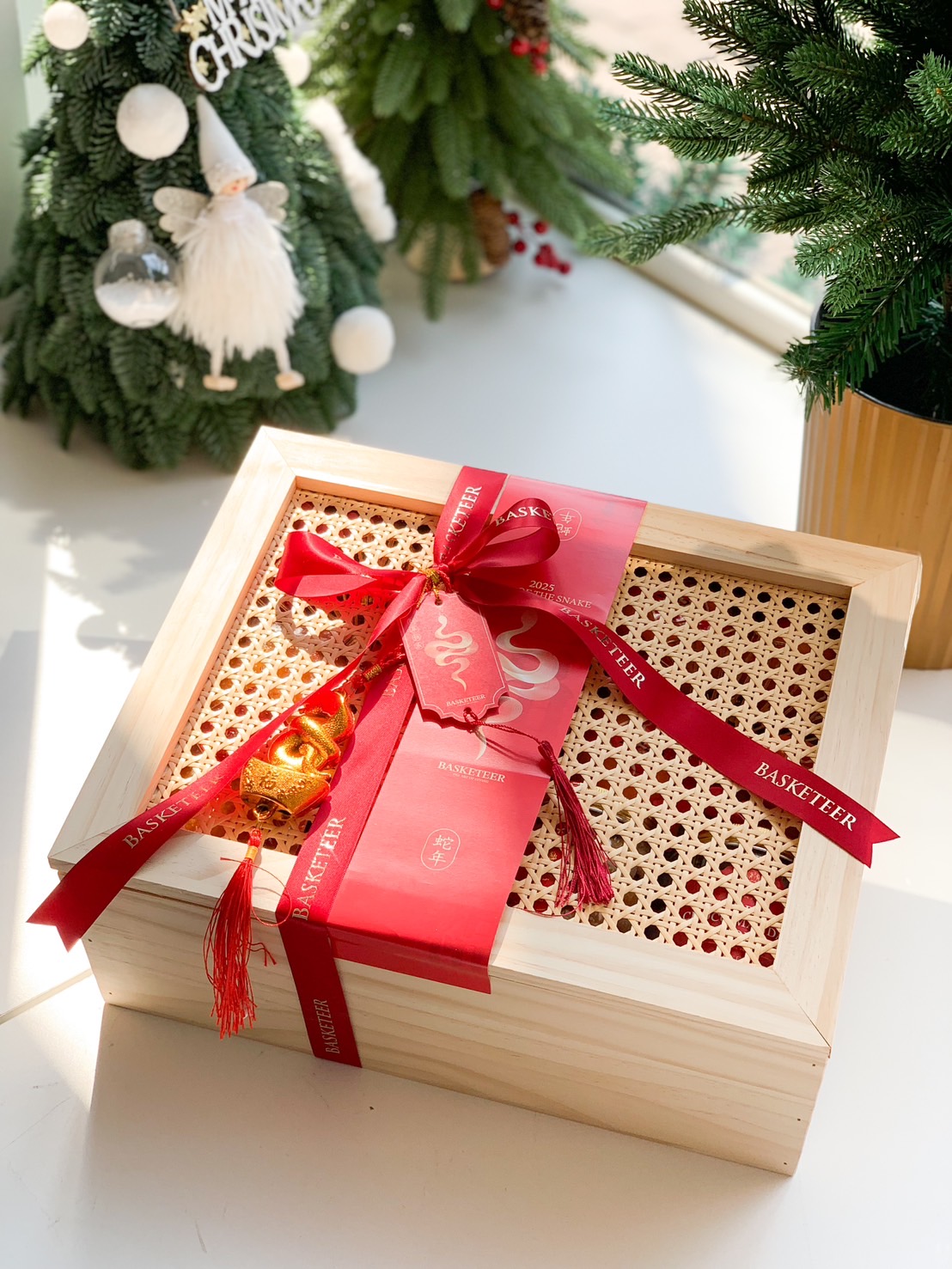 A wooden gift box filled with Chinese New Year items, including oranges, gold coins, a tin of butter cookies, candies, nuts, a jar of cookies, and a Lindt chocolate box. The box is set against a red background with traditional Chinese decorations.