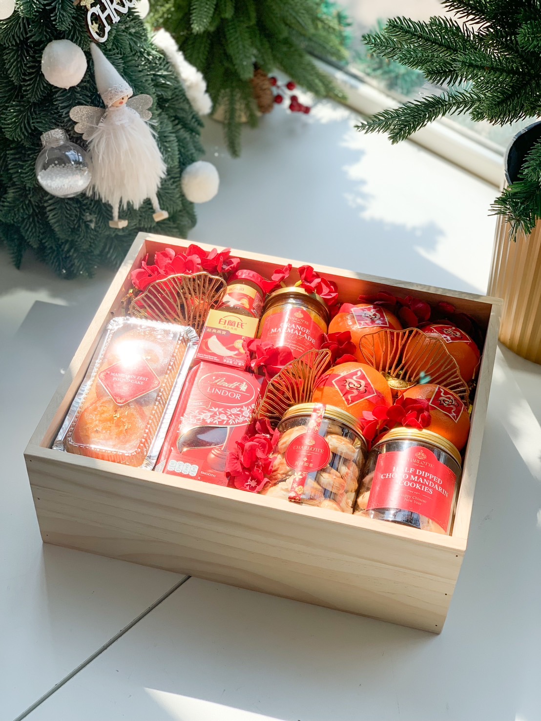 A wooden gift box filled with Chinese New Year items, including oranges, gold coins, a tin of butter cookies, candies, nuts, a jar of cookies, and a Lindt chocolate box. The box is set against a red background with traditional Chinese decorations.