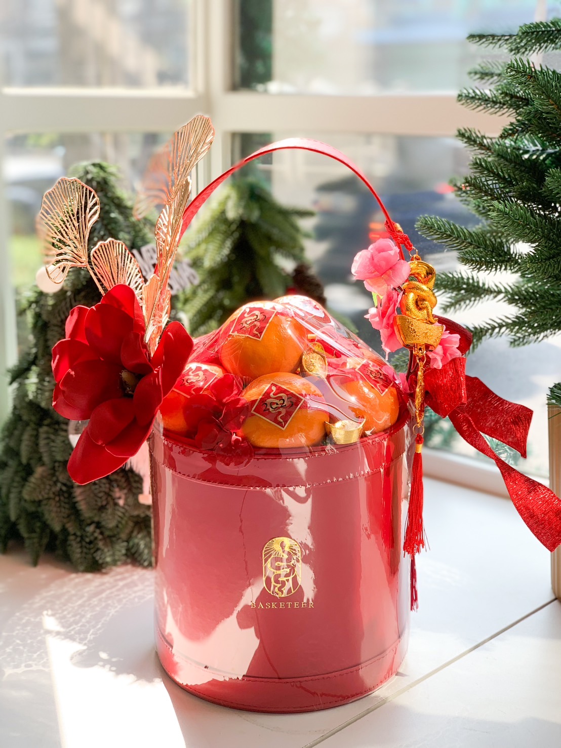 A red decorative basket filled with oranges, artificial flowers, and golden fan ornaments against a red background. The basket features a large red flower and small white blossoms. The backdrop includes folded red paper fans, enhancing the festive arrangement.