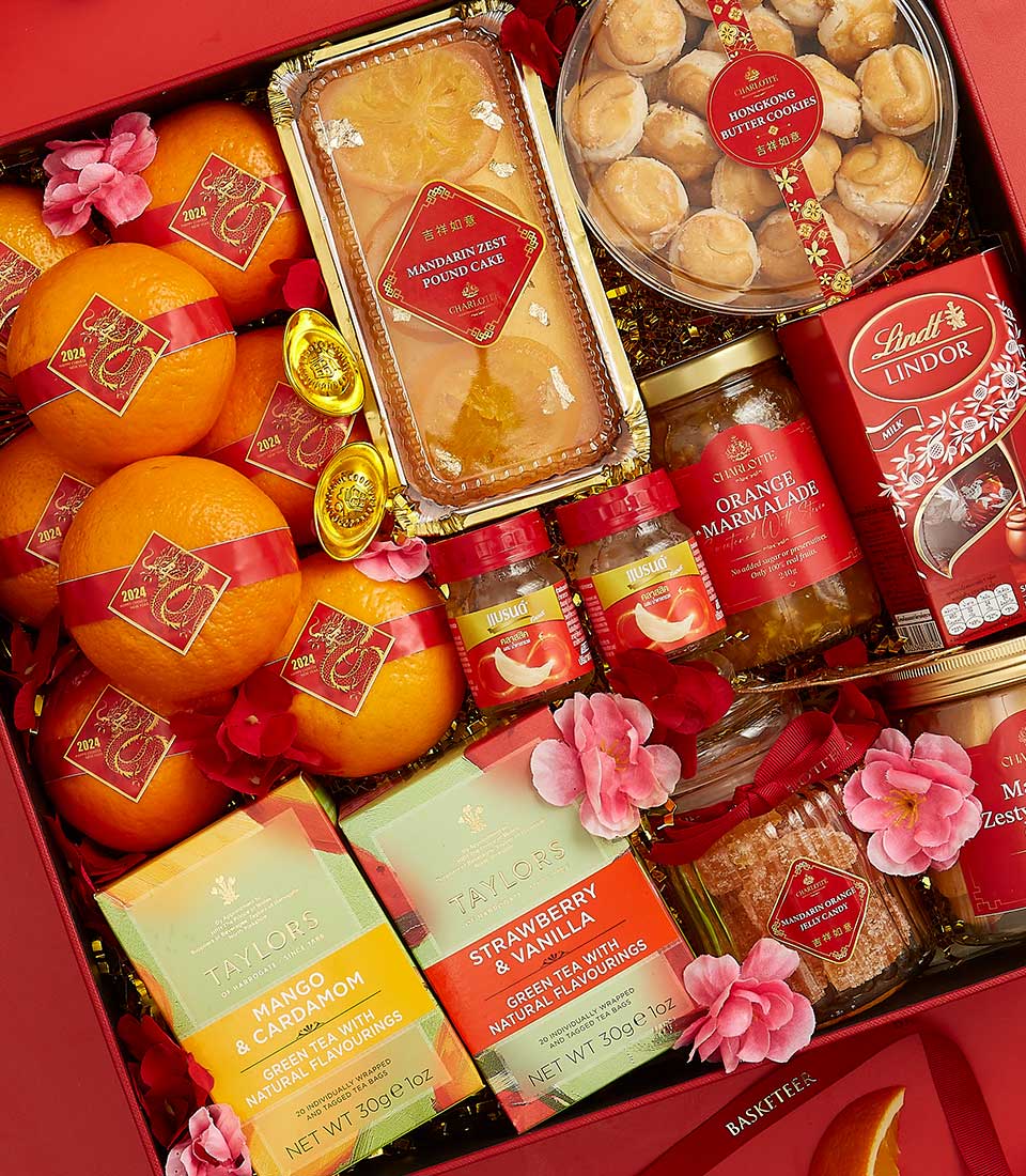 A gift basket filled with a variety of items including oranges, mango and cardamom tea, strawberry and vanilla tea, orange marmalade, a pound cake with orange zest, Lindt Lindor chocolates, and a box of round shortbread cookies. The basket is adorned with pink flowers.