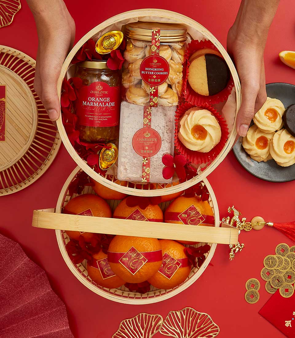 A person holding an open circular basket containing a jar of orange marmalade, cookies, bath salts, and red flowers. Another basket below holds several oranges. The setup is on a red surface with additional cookies and festive decorations around.