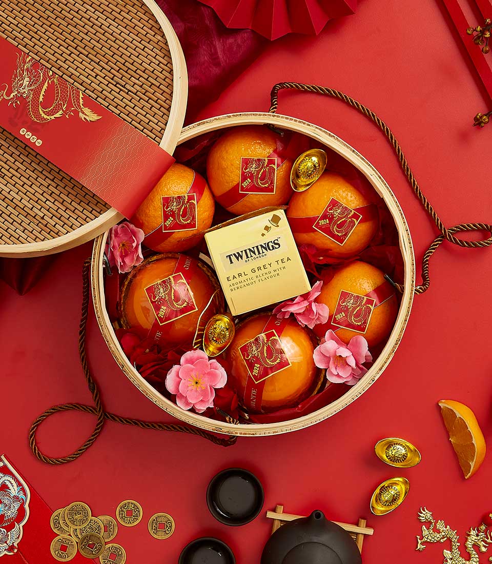 A round bamboo basket filled with oranges, pink flowers, golden ingots, and a box of Twinings Earl Grey tea. The basket lid, decorated with a dragon illustration, leans to the side on a red background adorned with various traditional items and decorations.