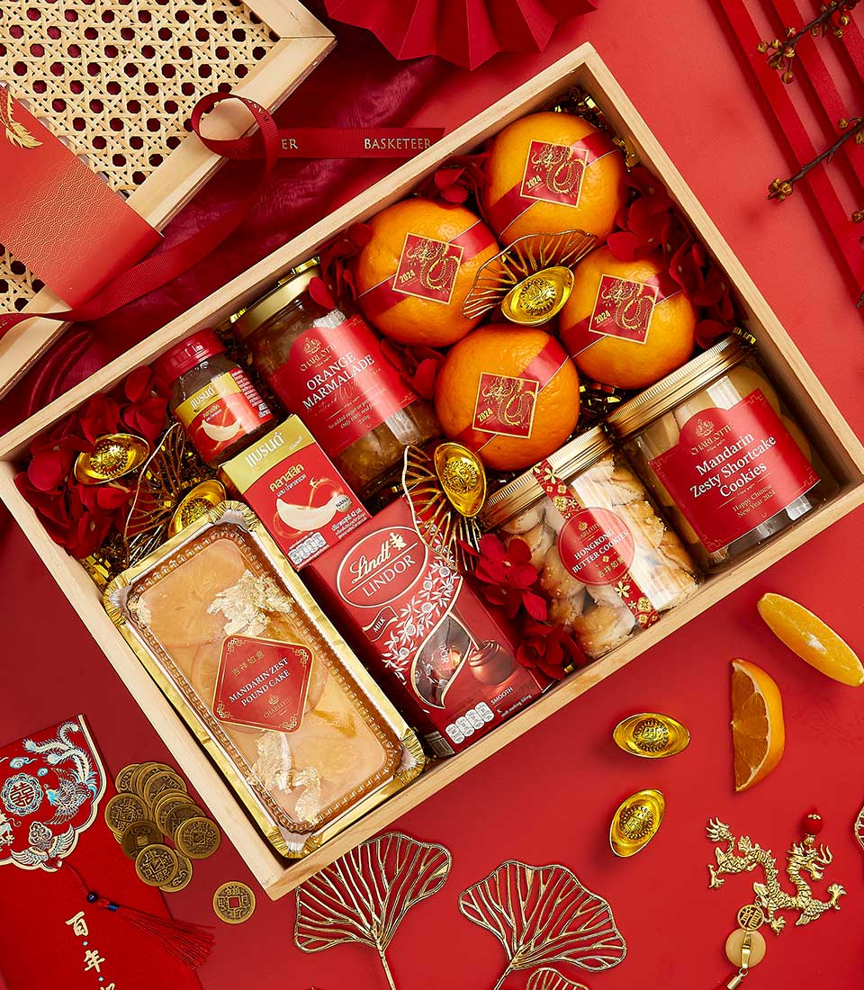 A wooden gift box filled with Chinese New Year items, including oranges, gold coins, a tin of butter cookies, candies, nuts, a jar of cookies, and a Lindt chocolate box. The box is set against a red background with traditional Chinese decorations.