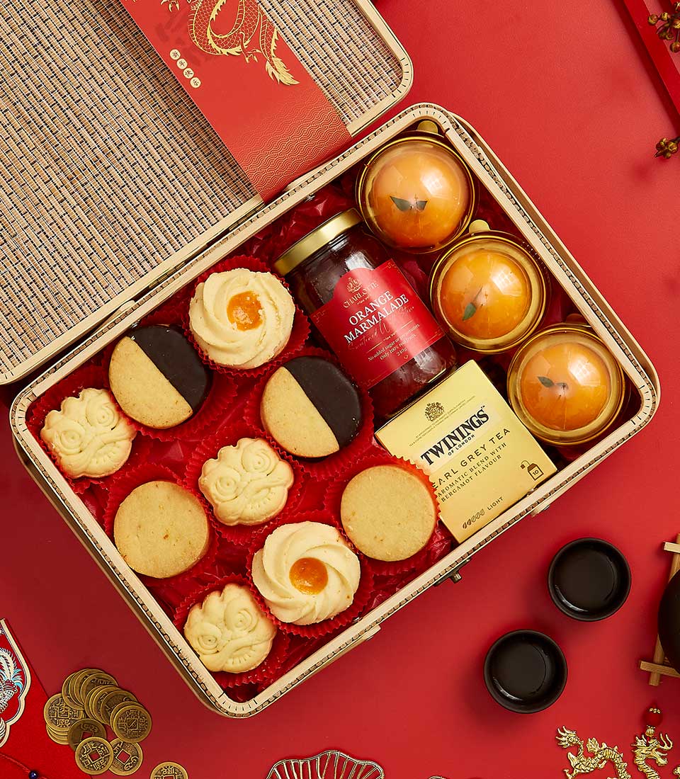 A decorative box contains various treats on a red background. Inside are cookies, three golden-wrapped round items, a jar of orange marmalade, a Twinings Earl Grey tea box, small round containers, and festive Chinese decorations.