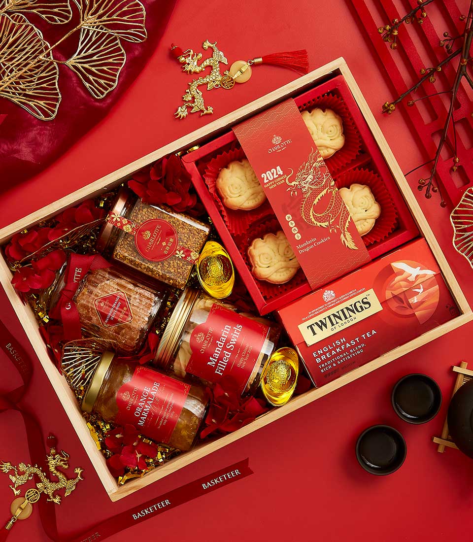 A festive gift box with various items arranged neatly on a red surface. The box includes jars of gourmet food, a Twinings English Breakfast Tea box, gold-colored coins, and a red packet with a dragon design. There are also decorative floral elements and two cups.