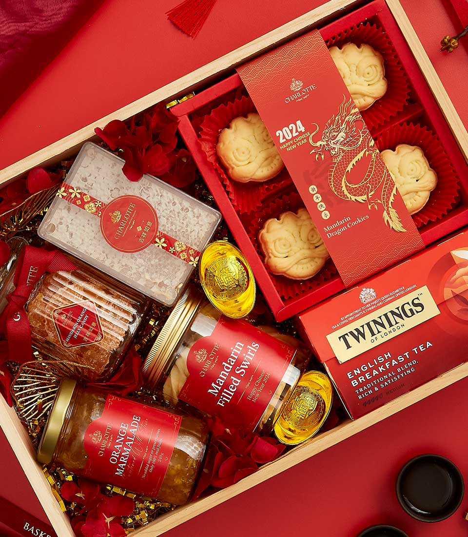 A wooden gift box with various items including Twinings English Breakfast tea, mandarin-flavored cookies, a jar of mandarin peel sugar, organic marmalade, a box with a gold dragon design, and two golden jars, surrounded by red fabric and decorations.