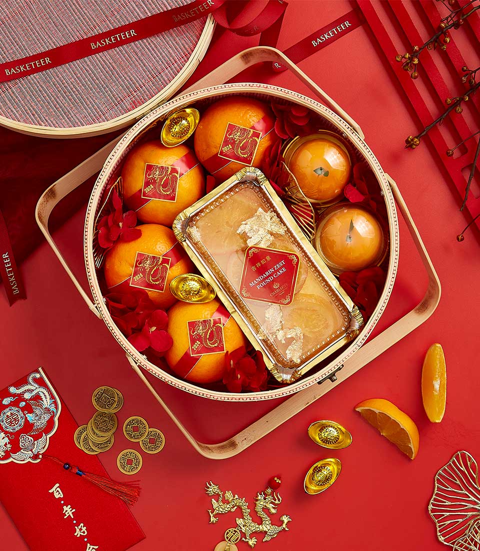 A festive basket containing oranges, a rectangular box with a gold-wrapped item, gold ingots, and red decorations sits open on a red surface. The basket lid and traditional Chinese ornaments, including red packets and coins, are displayed beside it.