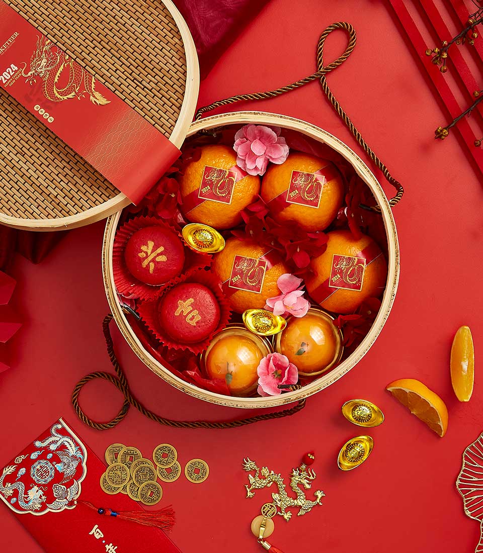 A festive arrangement for Lunar New Year featuring a round basket containing oranges, red packets, and gold ingots. The basket is surrounded by red envelopes, Chinese coins, decorations, and flowers, all set against a vibrant red background.