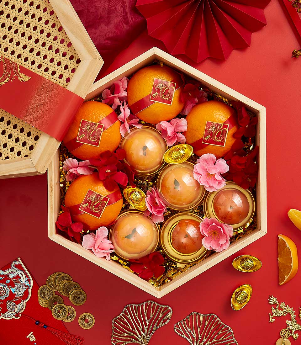 A festive hexagonal wooden gift box containing oranges, gold-wrapped spheres, and decorative flowers. Surrounding the box are gold ingots, artificial red foliage, a red pocket, and gold coins, all arranged on a red background for a celebratory occasion.
