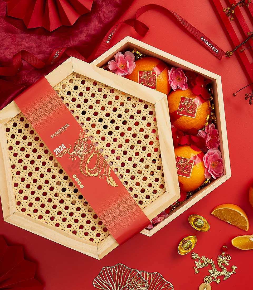 A festive Chinese New Year gift box is open, revealing three oranges adorned with gold-foil stickers and surrounded by pink flowers. The hexagonal box features a red cover with a dragon design and 