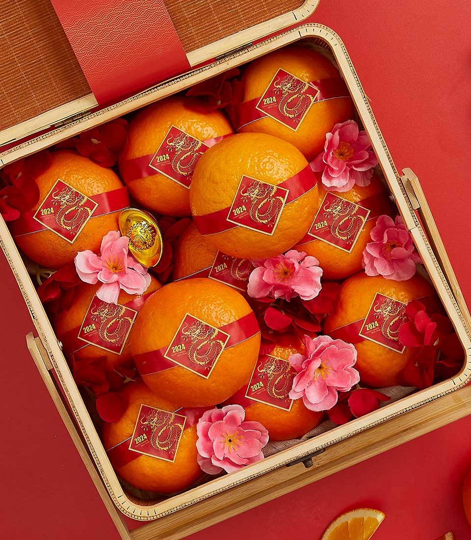 A decorative wooden CNY Mandarin Blessings Basket with oranges adorned with red ribbons and gold-embossed 2024 tags. The arrangement includes pink and red flowers, and a small golden ingot nestled among the oranges, all set against a red background.